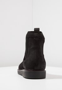 Bottines en cuir suédé noir avec col en tricot élastique, languette à l'arrière et semelle en caoutchouc épaisse sur une surface blanche.