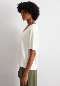 Cream V-neck T-shirt with short sleeves, made of soft fabric, featuring a relaxed fit and a straight hem, paired with olive green pants.