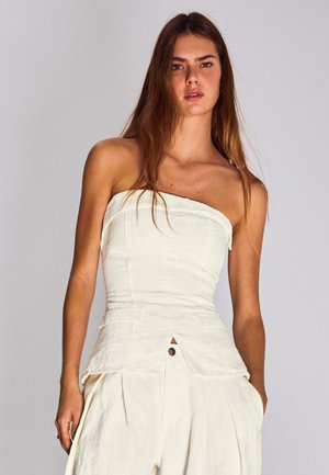 Hosszú barna hajú nő, fehér pántnélküli felsőt és hozzáillő, magas derekú nadrágot visel, sima világosszürke háttér előtt áll.