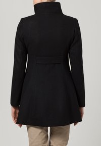 Svart, figursydd kappa med hög krage, långa ärmar och en något utsvängd fåll. Tillverkad av ull, med en slät yta och minimal detaljering.