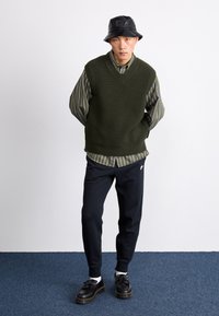 Vesta tricotată supradimensionată, de culoare verde închis, purtată peste o cămașă cu dungi, pălărie neagră tip bucket, pantaloni închiși la culoare și pantofi negri cu detalii din franjuri.