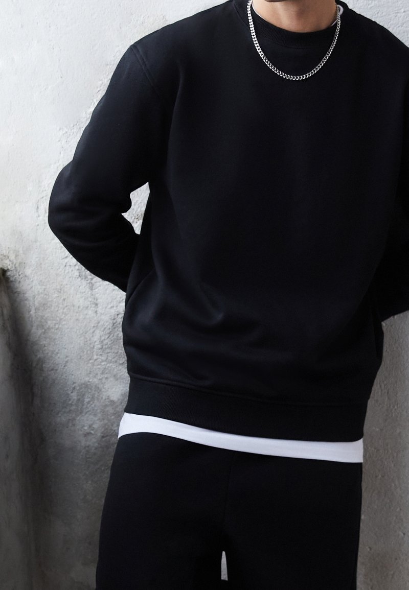 Personne portant une chemise à manches longues noire, un sous-vêtement blanc, un pantalon noir et un collier en chaîne argentée devant un mur clair texturé.