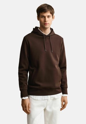 Jeune homme portant un sweat à capuche marron foncé par-dessus une chemise à rayures blanches avec un pantalon blanc, debout devant un fond uni clair.
