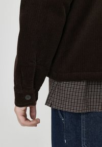 Veste en velours côtelé marron avec une texture côtelée, dotée d'un poignet à boutons. En dessous, une chemise à carreaux marron et blanche est visible.