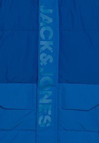 Blå vadderad jacka med en glänsande yta, stora frampocket och en vertikal logotryck i en ljusare blå nyans nedtill i mitten.