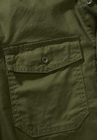 Tela de algodón verde oliva, con un solo bolsillo en el pecho con cierre de botón, detalle de costuras visibles y textura suave.