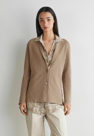 Frau trägt beige gerippten Cardigan über gemustertem Hemd mit hellbeiger Hose, steht vor einer einfachen weißen Wand.