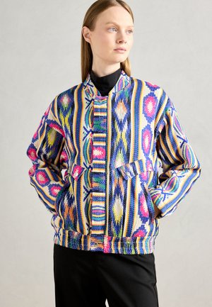 Donna che indossa un giacca vibrante a motivi multicolori con design a rombi e strisce, in piedi con le mani nelle tasche, che guarda di lato.