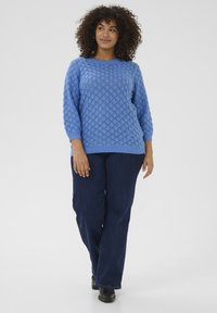 Blå strikket sweater med diamantmønster og ribbede manchetter, parret med mørkeblå lige ben jeans og sorte sko på en neutral baggrund.