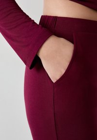 Bordeaux langesleeve top met fitted silhouet. Close-up toont een hand in een zijzak, wat het zachte materiaal en het naadloze ontwerp accentueert.