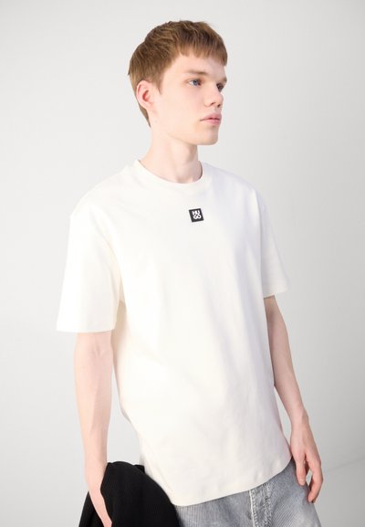 Кремаво-бял памучен T-shirt с къси ръкави и закръглен деколте. С квадратно черно лого на марката на гърдите. Обикновен дизайн, свободна кройка.