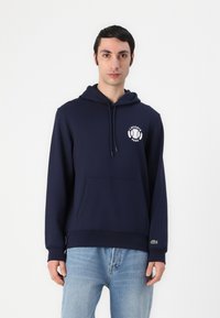 Felpa blu navy realizzata in tessuto morbido, con tasca frontale, cappuccio regolabile con coulisse e logo Lacoste bianco con design di alloro sul petto.