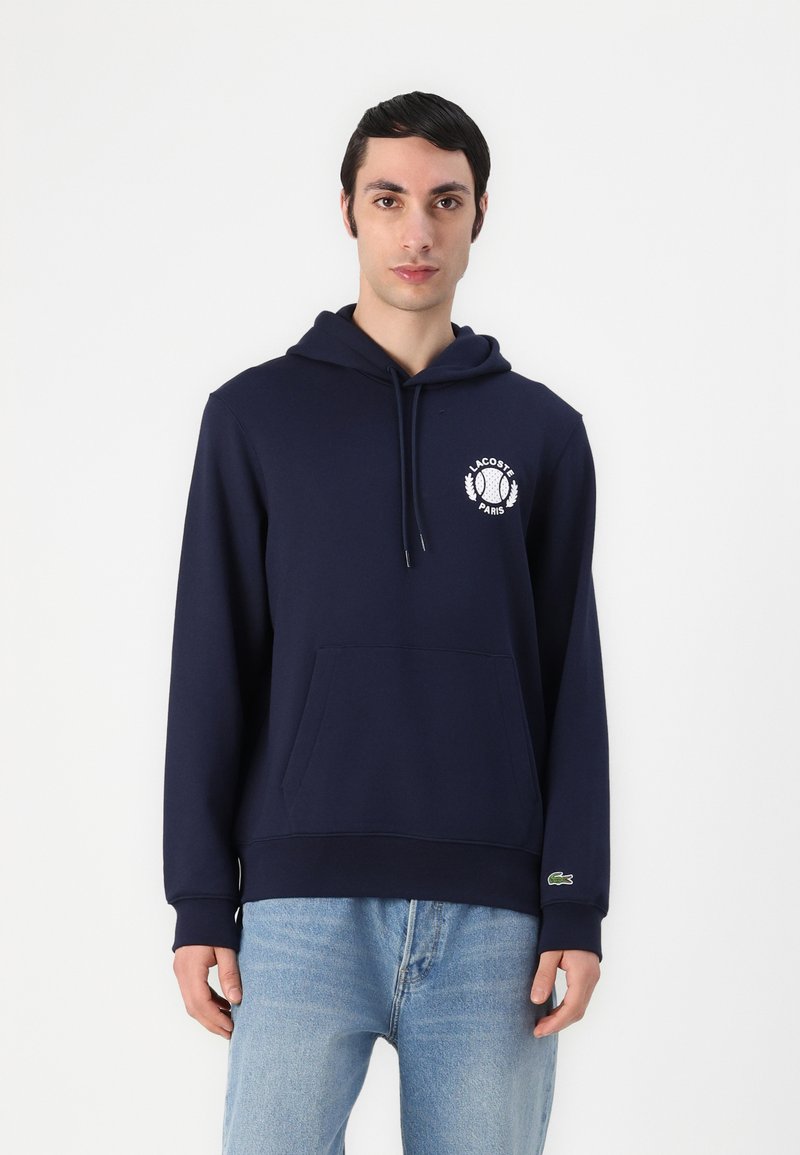 Felpa blu navy realizzata in tessuto morbido, con tasca frontale, cappuccio regolabile con coulisse e logo Lacoste bianco con design di alloro sul petto.