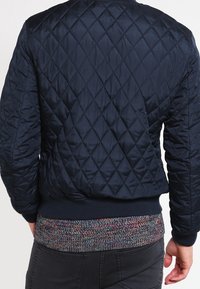 Chaqueta bomber acolchada de color marino con patrón de diamantes, dobladillo multicolor acanalado y puños ajustados, hecha de material ligero.