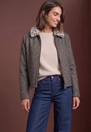 Une femme souriante porte une veste grise zippée à col en fausse fourrure, un pull crème et un jean bleu foncé, sur un fond marron.