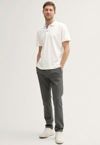 Weißes Poloshirt mit einem schwarz-weißen Kragen, kombiniert mit grauen Hosen und weißen Sneakers. Glatte Textur, lässige Passform und Knopfdetails.