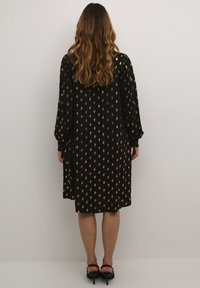 Črna obleka z zlatimi pikami, dolgi rokavi in ohlapen kroj. Teksturirana tkanina z elastičnimi manšetami. Obleka do kolen.