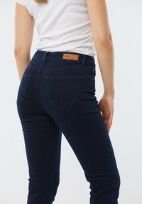 Jeans en denim bleu foncé avec une coupe ajustée, comportant une patch en cuir sur la taille arrière, deux poches arrière et des détails de couture subtils.