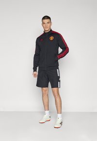 Sort atletisk jakke med røde striber og Manchester United-logo, parret med sorte shorts med hvide striber og hvide fodboldstøvler.
