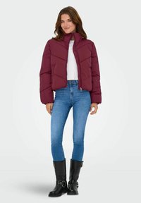 Vinski skraćeni puffer kaput s visokim ovratnikom i chevron šavovima, uparen s uskim plavim trapericama i crnim čizmama do koljena s kopčama.