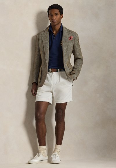 Καφέ σακάκι με σχέδιο houndstooth πάνω από μπλε πόλο, λευκό σορτς, καφέ ζώνη, λευκά αθλητικά παπούτσια και μπεζ κάλτσες. Μαντηλάκι τσέπης στο σακάκι.