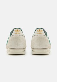 Adidas-skor med övre i ljusbeige mocka, gröna detaljer och en vit häldel med logotypen. Slät yta och rundad form.