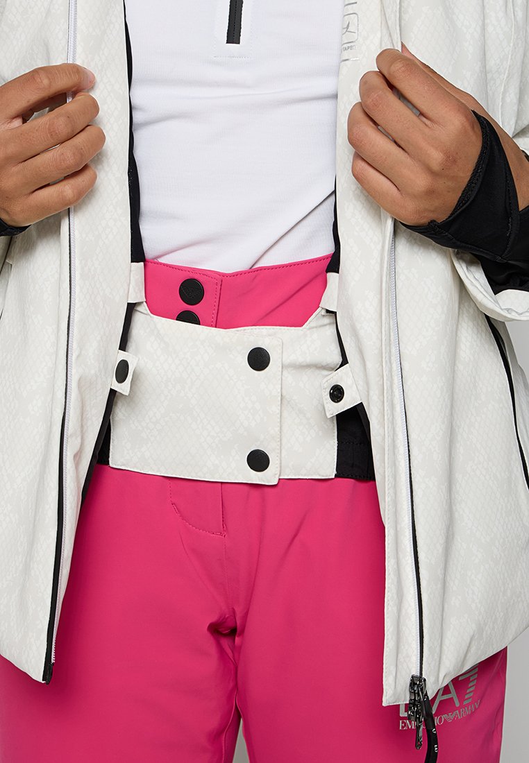 Weiße wasserdichte Jacke mit strukturiertem Finish und schwarzen Akzenten, kombiniert mit leuchtend pinkfarbener Skihose, die mit Schnallen- und Druckknopfdetails versehen ist.