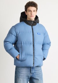 Blue insulated puffer jacket with a black hood, front zipper, and logo patch. Features quilted design, side pockets, and a relaxed fit.