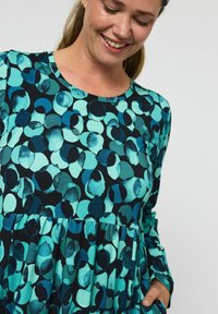 Šaty s dlhým rukávom, ktoré majú abstraktný vzor modrozelených a čiernych kruhov; mäkká tkanina s naberaným pásom a okrúhlym výstrihom.