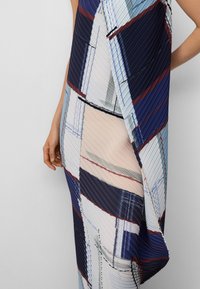 Vrouw in een geplooide, meerkleurige diagonale gestreepte jurk met marineblauwe, lichtblauwe, bordeauxrode en beige tinten, staande tegen een effen muur.
