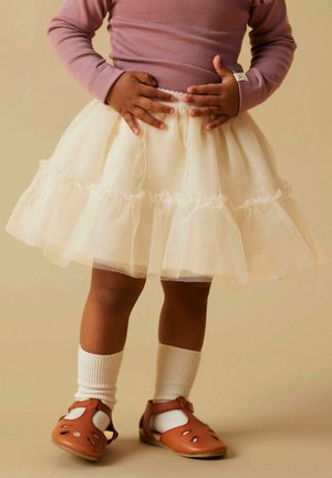 Weißer Tüllrock mit rüschengeschichteten Lagen, getragen über weißen gerippten Socken und braunen Sandalen mit Ausschnitten. Das Oberteil ist helllila.