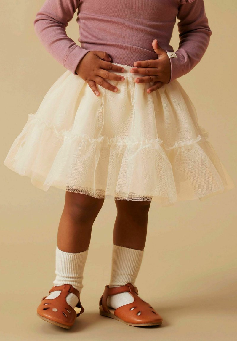 Bela tilna krila z nabrano plastjo, nosena čez bele rebraste nogavice in rjave sandale s izrezljanimi detajli. Zgornji del je svetlo vijoličen.