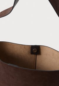 Hnědá semišová taška s hladkým vnitřním podšívkou, vybavená magnetickým uzávěrem a mírně zakřiveným horním okrajem. Jednoduchý a funkční design.