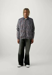 Persona con cabello trenzado hasta los hombros, vistiendo una chaqueta gris de patrón con cremallera, pantalones amplios de mezclilla negra deslavados y zapatillas blancas.