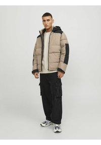 Giacca imbottita beige con dettagli neri, con chiusura a zip e cappuccio. Indossata sopra una maglietta chiara e abbinata a pantaloni cargo neri.