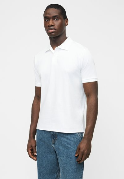 Homme portant un polo blanc à manches courtes et un jean bleu, debout devant un fond blanc uni.