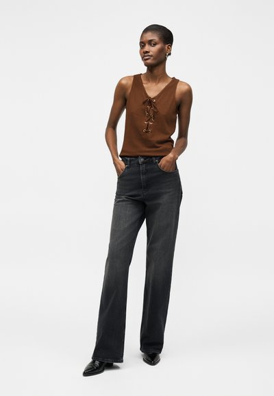 Γυναίκα που φοράει καφέ αμάνικη μπλούζα με κορδόνια μπροστά, μαύρο τζιν με ψηλή μέση και φαρδιά πόδια, και μαύρα μυτερά παπούτσια, στέκεται με τα χέρια στις τσέπες.