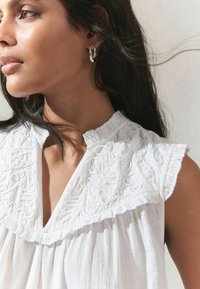 Haut blanc texturé avec des détails brodés, col volanté et manches courtes. Des boucles d'oreilles en argent complètent la tenue.