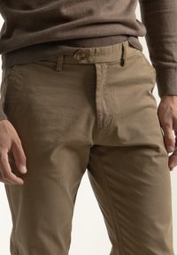 Pantalones de algodón marrones con un corte ajustado, que cuentan con cierre de botones, bolsillos laterales y una textura suave. Detalles de costura visibles.
