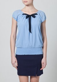 Blusa de manga corta azul claro con escote fruncido y lazo de cinta azul marino; cuenta con una cintura fruncida texturizada. Combinada con una falda azul marino.