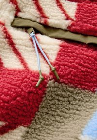 Tissu en polaire rouge, crème et marron avec un motif géométrique. Présente une bordure verte et des cordons de serrage bleus pour l'ajustement.