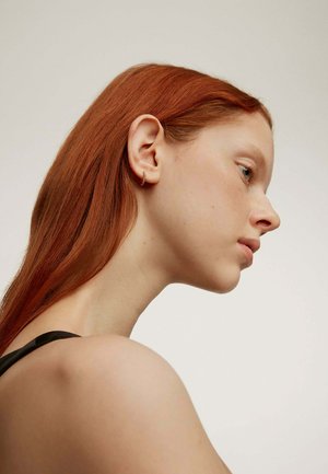 Pendiente de aro dorado en una modelo con cabello rojo largo, mostrado de perfil contra un fondo neutro, con una textura suave y un diseño sutil.