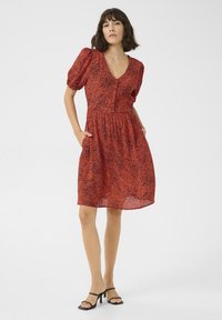 Robe rouge avec un motif tacheté noir, décolleté en V, manches bouffantes et poches. Fabriquée en tissu léger, longueur genou, avec une taille froncée.