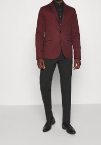 Burgundowa marynarka o teksturowanym wykończeniu, zapinana na jeden guzik, z dwoma przednimi kieszeniami, w połączeniu z czarnymi spodniami i formalnymi butami.