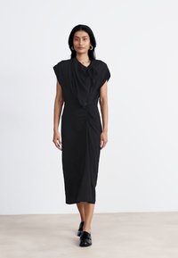 Robe midi noire ajustée avec des manches courtes, détails froncés à la taille et texture lisse. Le mannequin porte des chaussures noires.
