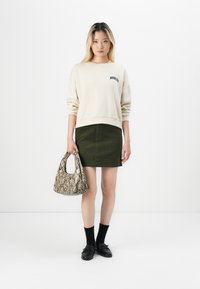 Sweatshirt crème avec le texte "BROOKLYN", jupe en velours côtelé vert, sac en cuir texturé effet peau de serpent, chaussettes noires et mocassins noirs.