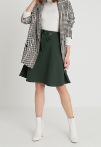 Jupe évasée verte avec un détail de taille à nouer, associée à un blazer à carreaux noir et blanc et des bottines blanches.