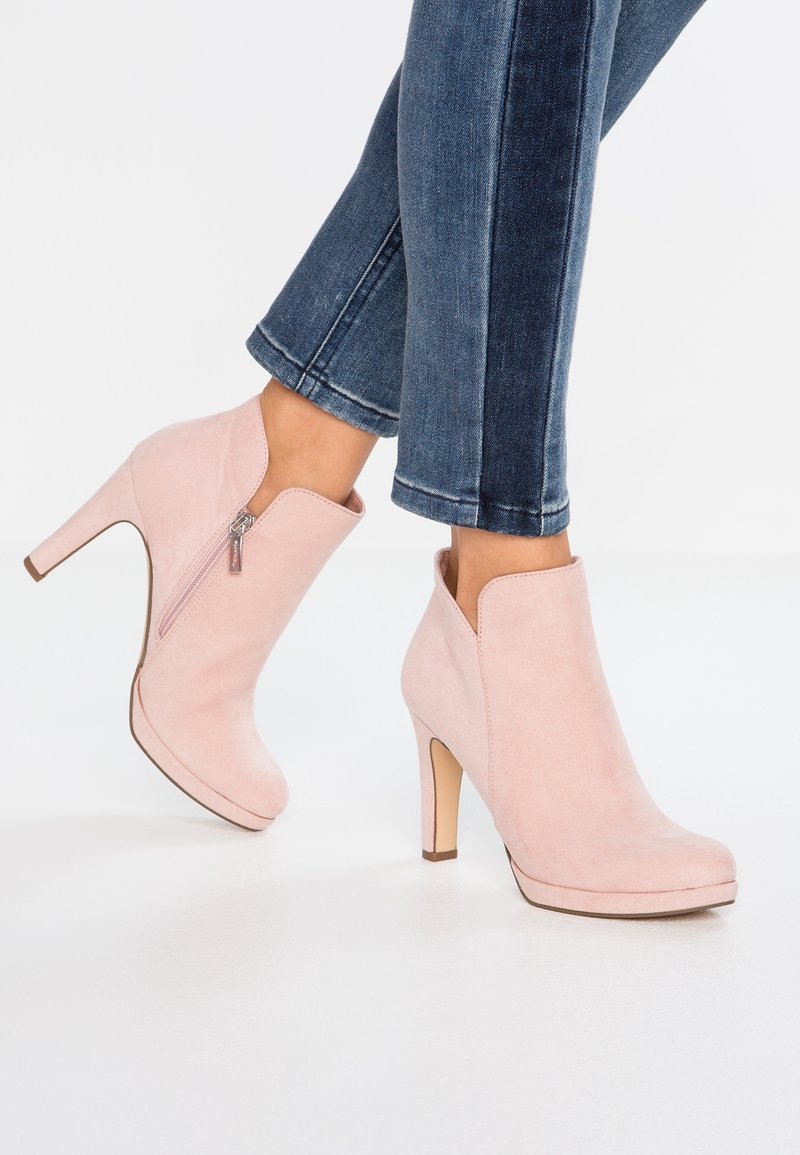 Pieds portant des bottines en daim rose clair avec des fermetures éclair latérales et des talons carrés, associées à un jean en denim bleu coupé au-dessus de la cheville.