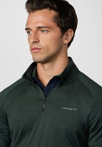 Dunkelgrüner Pullover mit Reißverschlusskragen, strukturierter Stoff und einem dezenten Logo auf der Brust. Darunter befinden sich ein navyblaues Hemd.