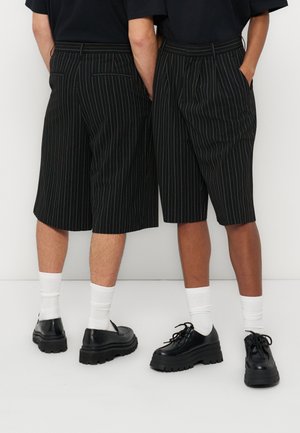 Twee mensen die zwarte kniebroeken met krijtstrepen, zwarte tops, witte sokken en stevige zwarte schoenen dragen, staand met handen in de zakken.